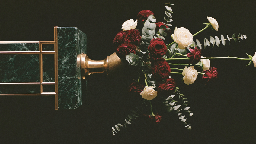 Skulpturale Blumenkomposition aus dunklen Ranunkeln und cremefarbenen Rosen auf einem Marmorsockel, inszeniert wie ein Ausstellungsstück