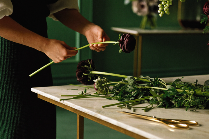 Hände setzen behutsam einen dunkelroten Ranunkelstiel in eine entstehende Blumenkomposition auf einem Marmortisch